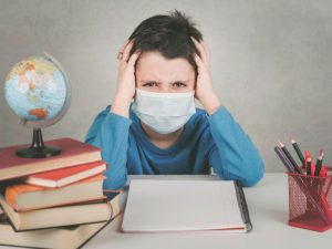 a young boy wearing a mask clasps his head in frustration next to school books