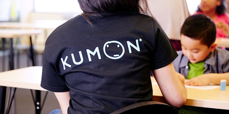 A Kumon instructor wearing a black shirt helping a student becuase to become a better writer you must read