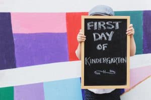 Child holding up "First Day of Kindergarten" sign in front of a colorful mural.