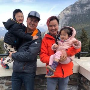 Jason and Rico stand in front of a mountain holding their kids