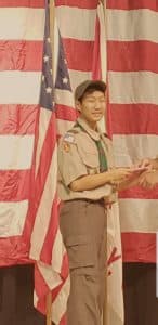 Ryan stands in front of an American flag in his Boy Scouts uniform