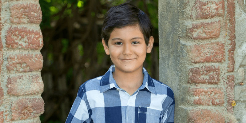 Yaseen stands against a brick wall wearing a blue plaid shirt