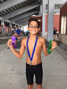 Nicholas stands in his swim uniform holding ribbons with a medal around his neck