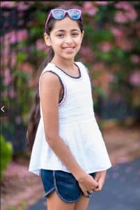 Asmita stands in a driveway with flowers behind her, wearing a white tank top and glasses on her head.