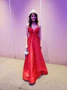 Khushi stands in a plain room wearing a red dress and tiara