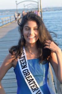Khushi stands on a pier wearing a blue dress and sash as the wind blows her hair