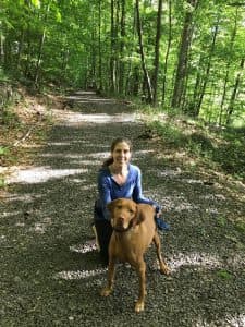 Lillian squats next to her dog on a path in the woods