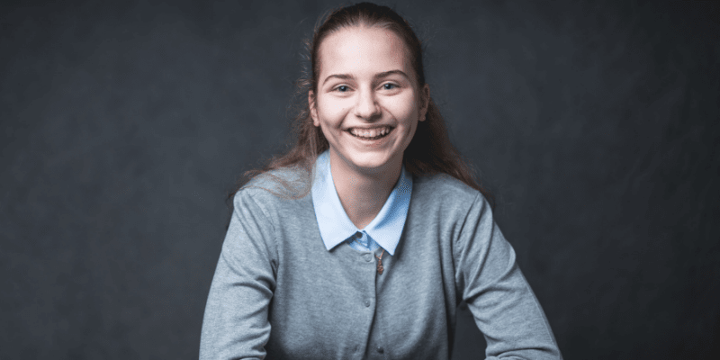 Sophia grins in front of a gray background, wearing a gray sweater over a blue collared shirt