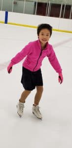 Alvina figure skates wearing a pink fleece top and black skirt