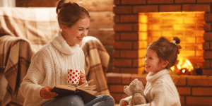 A other and daughter sit in front of a fireplace wearing matching white sweaters and reading