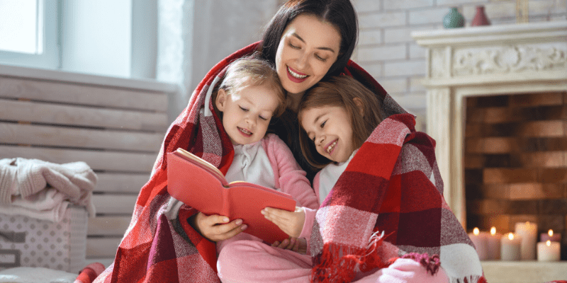 A mother sits in front of the fireplace with her two daughters wrapped in a blanket and reading snow stories