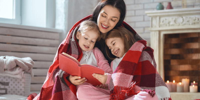 A mother sits in front of the fireplace with her two daughters wrapped in a blanket and reading a book