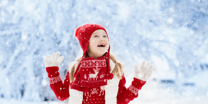 Young blonde girl wearing red jacket, scarf, and hat. Her white gloves are extended waiting for the snow to fall on them.