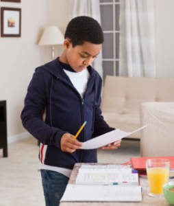 Boy wearing navy blue jacket with pen and paper in hand reviewing his assignments in his living room.