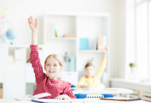 Girl with red and white checkered shirt raises hand while smiling in class