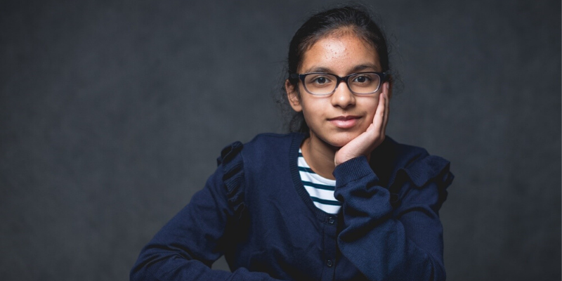 Young girl wearing striped shirt and a navy blue cardigan. She has one hand on her face as if she's thinking while sporting her black square glasses.
