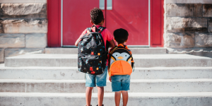 Two young boys, one taller than the other, wearing backpacks standing in front of the steps to the red door that leads to their school entrance