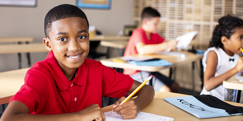 Boy in red polo shirt holding a pencil and working on his Kumon worksheets while smiling at the camera.