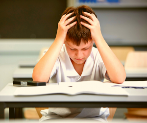 Boy in a classroom wearing a white shirt with both of his hands on his head while reading.