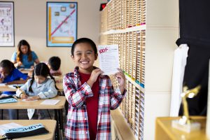 Young boy in a plaid shirt is holding up his Kumon worksheet with 100% grade.
