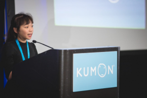 Student speaker behind a Kumon podium wearing a black shirt and blue Kumon lanyard around her neck
