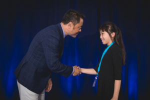 Young girl with black shirt and blue Kumon lanyard shakes hands with KNA President Mino