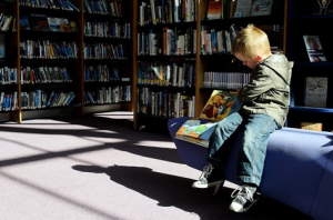 Blonde boy sitting at a private section of a library reaching over to his right side while he flips page of a book.