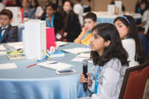 student holds a mic as she speaks from her round table full of students