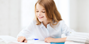 Girl in white shirt smiling and writing notes in a blue pen pointing at something in her notebook.