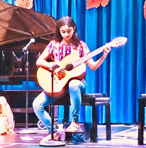 young lady playing the guitar sitting on black bench on stage with blue curtains