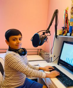 A young boy with headphones around his neck sits in front of a computer with his hands on the keyboard, speaking into a microphone